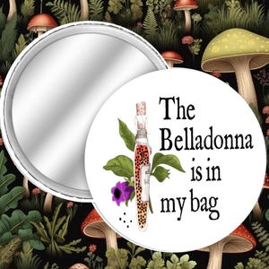 May include: A round compact mirror with a white design featuring a bottle with a cork, green leaves, and the text "The Belladonna is in my bag." The background has a whimsical mushroom and plant pattern.