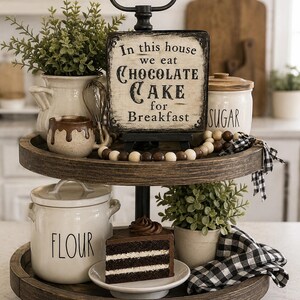 May include: A two-tiered wooden tray featuring a sign that reads In this house we eat CHOCOLATE CAKE for Breakfast. The tray holds a white flour canister, a sugar canister, a slice of chocolate cake on a plate, a small plant, and a mug.