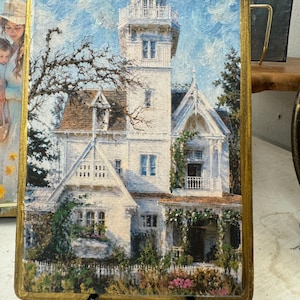 May include: A rectangular wall art piece featuring an oil painting of a white multi-story house with a prominent tower, surrounded by green foliage and a white picket fence, all enclosed within a textured gold-painted wooden frame.