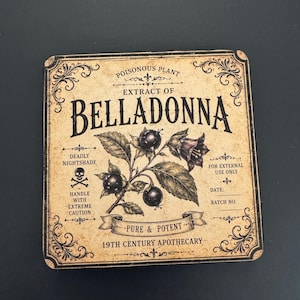 May include: A square coaster with a vintage-style beige label featuring the text "EXTRACT OF BELLADONNA" and "19TH CENTURY APOTHECARY." The design includes a botanical illustration of dark berries, a skull and crossbones icon, and decorative borders.