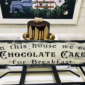 May include: A wooden sign with black lettering that reads "In this house we eat chocolate cake for breakfast". A small chocolate cake with a chocolate drizzle is on a plate in front of the sign.
