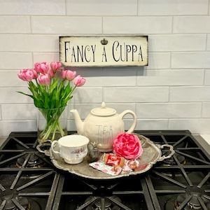 May include: A white teapot with the word "TEA" on it, a white teacup, pink flowers in a glass vase, and a silver tray with pink and white candy on a black stovetop. A wooden sign with the text "FANCY A CUPPA" is hanging on a white tiled wall.