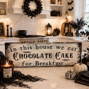 May include: A Halloween-themed kitchen scene with a wooden sign that reads "In this house we eat CHOCOLATE CAKE for Breakfast." Decorative pumpkins, candles, and a witch's hat are arranged on the counter. A "Trick or Treat" pumpkin is also visible.