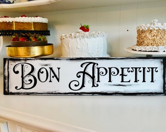 Puede incluir: Un letrero de madera blanco con letras negras que dice "Bon Appetit" en una fuente de letra decorativa. El letrero tiene un aspecto desgastado y vintage con pintura negra alrededor de los bordes.