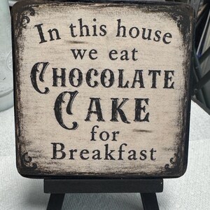 May include: A square wooden sign with a distressed cream finish and black edges, featuring the black text In this house we eat CHOCOLATE CAKE for Breakfast. The sign is displayed on a small black wooden easel stand with four short legs.
