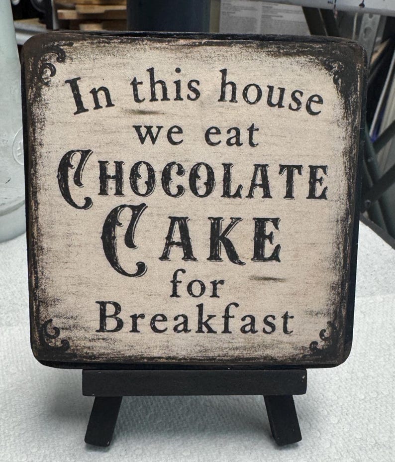 May include: A square wooden sign with a distressed cream finish and black edges, featuring the black text In this house we eat CHOCOLATE CAKE for Breakfast. The sign is displayed on a small black wooden easel stand with four short legs.