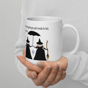 May include: White ceramic mug with a black and white illustration of two witches holding umbrellas and brooms. The text on the mug reads "If you have a garden gate; plant lavender for luck; if you can."