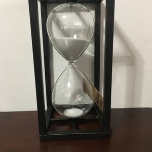 May include: An hourglass with white sand inside a clear glass bulb, encased in a black metal frame. The hourglass has a tag that reads "Metal Hourglass". The frame is rectangular and sits on a dark wooden surface.
