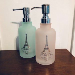 May include: Two frosted glass soap dispensers, one mint green and one pale pink, with silver pump tops. Each bottle features a silver Eiffel Tower design with the text "PARIS RUE DES ROSIERS 1918".