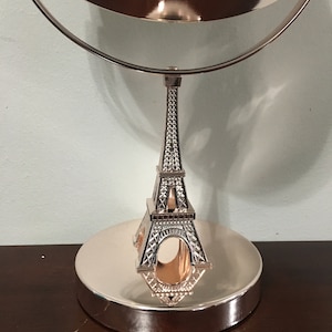 May include: Rose gold vanity mirror with a double-sided design. The base features a circular shape, supporting a detailed Eiffel Tower structure. The mirror's frame is also rose gold, creating a cohesive and elegant aesthetic.