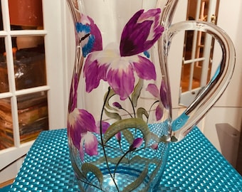 May include: A clear glass pitcher with a curved handle, adorned with hand-painted purple and white floral designs. The pitcher features a wide mouth and a slightly flared body, perfect for serving beverages. The floral design includes green leaves and stems.