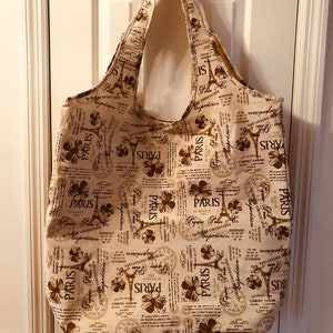 May include: Beige tote bag with a repeating pattern of the Eiffel Tower, four-leaf clovers, and the word "Paris" in brown. The bag has a wide, open top and long straps for carrying over the shoulder.