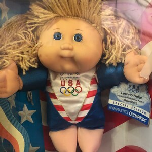 May include: A Cabbage Patch Kid doll dressed in a blue and red gymnastics leotard with the USA Olympic logo and the words "Official Team Mascot" on the front. The doll has blonde hair and blue eyes. The doll is packaged in a box with a red, white, and blue American flag design. The box also includes the text "USA Leanna Helen was born in the Cabbage Patch on December 1 SPECIAL EDITION GYMNASTICS OLYMPIKIDS."