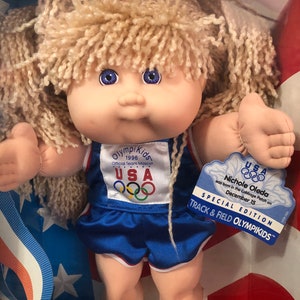 May include: A Cabbage Patch Kids doll dressed in a blue and white track and field uniform with the 1996 Olympic mascot logo. The doll has blonde hair and blue eyes. The doll is holding a thumbs up with both hands. The doll is in a box with a red, white, and blue background. The box also has a special edition tag that says "USA Nichole Oleda was born in the Cabbage Patch on December 15. Special Edition Track & Field Olympikids."