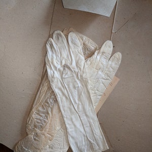 May include: A pair of off-white gloves, likely vintage, are laid out on a cardboard surface. The gloves are long, reaching past the wrist, and appear to be made of a delicate material. The gloves are in good condition.