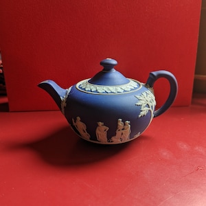May include: A blue ceramic teapot with a white bas-relief design. The teapot features a curved spout, a rounded body, and a handle. The lid is topped with a small knob. The design includes figures and foliage. The teapot is set against a red background.