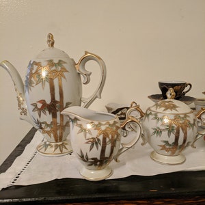 May include: A vintage tea set with a white background and gold bamboo design. The set includes a teapot, creamer, sugar bowl, and teacups. The teapot has a curved handle and a decorative lid. The set is displayed on a white cloth.