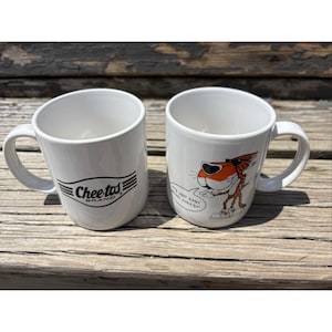 May include: Two white ceramic mugs. The left mug features the black Cheetos Brand logo with horizontal lines. The right mug displays a graphic of the Cheetos cheetah character wearing sunglasses with a speech bubble that reads IT'S NOT EASY BEING CHEESY.