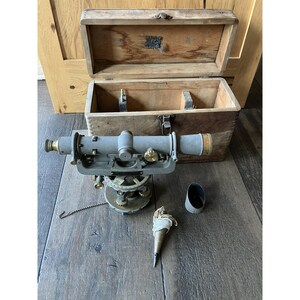 May include: An antique transit level with a wooden storage box. The level is gray with brass accents and sits on a brass base. A plumb bob, a sighting tube, and a chain are also present. The open wooden box is light brown.