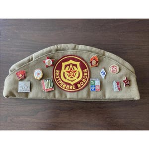 May include: A tan military cap with multiple pins and badges. The cap has a rounded shape and is made of a textured fabric. The pins and badges feature various designs, including stars, flames, and text in a foreign language. The cap is displayed on a wooden surface.