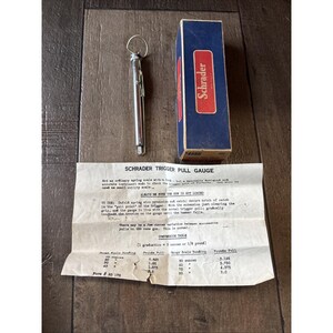 May include: A Schrader trigger pull gauge set, including a silver metal gauge, a blue and red box, and an instruction sheet. The gauge measures trigger pull in pounds. The box has the number 4556.