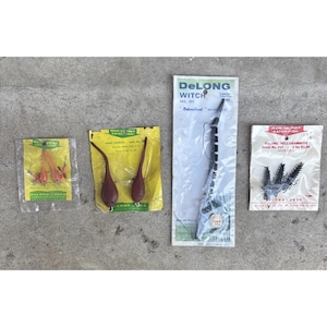 May include: Four packages of DeLong fishing lures. The first is a small yellow pack with two pink lures. The second is a yellow pack with two brown tadpole lures. The third is a long white pack with a black segmented lure. The fourth is a white pack with three black hellgrammite lures.