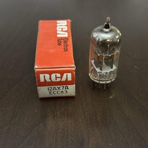 May include: An RCA Electron Tube, model 12AX7A ECC83, sits next to its original red box. The tube is clear with internal components visible. The box has the RCA logo and tube information printed in white.