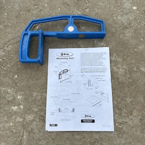 Puede incluir: Una herramienta de montaje Kreg de plástico azul con un mango y dos imanes circulares, acompañada de un manual de instrucciones blanco con diagramas y texto en negro que detalla el proceso de instalación de guías de cajón.