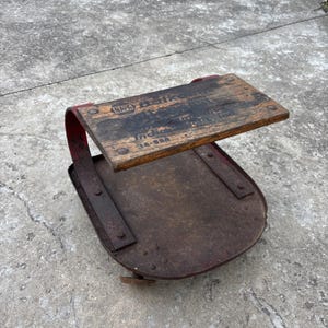 May include: A vintage metal and wood creeper seat with a weathered red finish. The rectangular wooden top features the NAPA logo and text including 'Indi' and '14-98A'. The base is a rounded metal plate with curved supports and small wheels.