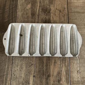 May include: A vintage, silver-colored cast iron cornbread mold with six individual corn-shaped cavities. The mold has a rectangular shape with a handle on one end and is set against a wooden surface.