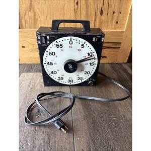 May include: A black square industrial timer with a white circular face featuring black and red numerals from 0 to 55. The device has a top handle, a central black knob, two small switches labeled Timer at Zero and Power, and a black electrical cord.