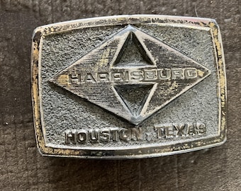 Puede incluir: Hebilla de cinturón rectangular plateada con el texto "HARRISBURG" dentro de un rombo. Debajo del rombo, el texto "HOUSTON, TEXAS" es visible. La hebilla tiene un aspecto desgastado y vintage.