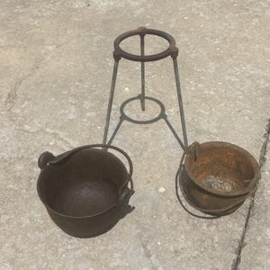 May include: Two small, dark brown cast iron pots with handles sit beside a metal tripod stand. The pots have a rounded shape and are likely for cooking or heating. The stand has a circular top and base.