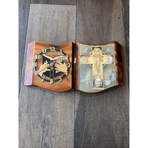 May include: A wooden wall clock shaped like an open book. The left side features a clock face with the word "Bible" and hands. The right side displays a religious image of a cross and figures. The clock is brown with a dark border.