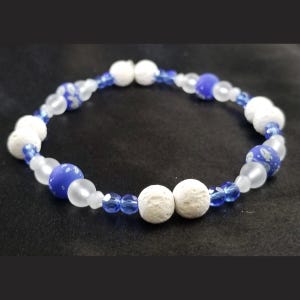 May include: A beaded bracelet featuring a repeating pattern of white porous lava beads, translucent frosted white beads, small faceted blue glass beads, and larger opaque blue beads with a white speckled pattern arranged in a circular design.
