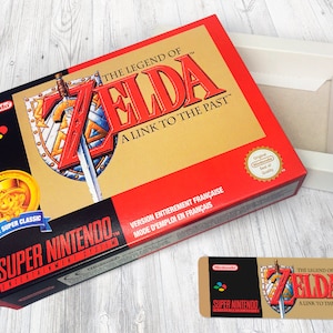 May include: A red and gold Super Nintendo game box for The Legend of Zelda: A Link to the Past. The box features a gold coin with a cartoon character wearing a hat and a sword and shield design. The text "Version Enti&egrave;rement Fran&ccedil;aise" is printed on the box.
