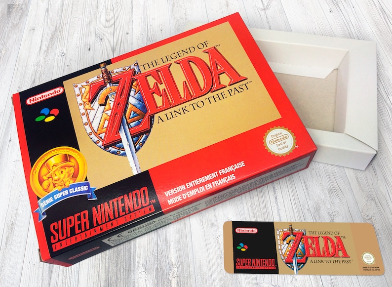 May include: A red and gold Super Nintendo game box for The Legend of Zelda: A Link to the Past. The box features a gold coin with a cartoon character wearing a hat and a sword and shield design. The text "Version Enti&egrave;rement Fran&ccedil;aise" is printed on the box.