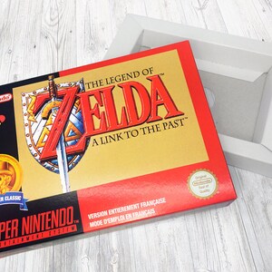 May include: A red and gold Super Nintendo game cartridge box for The Legend of Zelda: A Link to the Past. The box features a sword and shield design with the game title and a Nintendo logo. The box is in French.