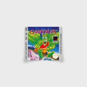 Puede incluir: Un cartucho de Game Boy para el juego "Gargoyles" con una etiqueta verde, roja y blanca. La etiqueta presenta un personaje de caricatura de gárgola con un atuendo rojo y blanco. El texto "THIS SIDE OUT/ CE CÔTÉ À L'EXTÉRIEUR" está impreso en el lado derecho de la etiqueta.