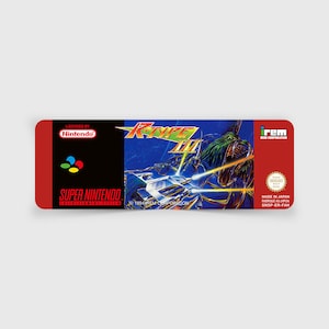May include: A Super Nintendo Entertainment System game cartridge for the game R-Type III: The Third Lightning. The cartridge is red and black with a colourful image of a spaceship and a monster on the front. The text "Licensed by Nintendo", "Super Nintendo", "1994 Irem Corporation", "irem", and "Made in Japan" is printed on the cartridge.