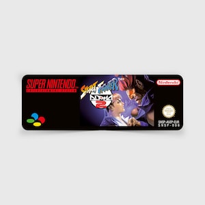 Może przedstawiać: Kaseta gry Super Nintendo Entertainment System do gry Street Fighter Alpha 2. Kaseta jest czarna z kolorową etykietą przedstawiającą logo i postacie z gry. Etykieta zawiera również tekst "SNSP-AUZP-EUR" i "SNSP-006".