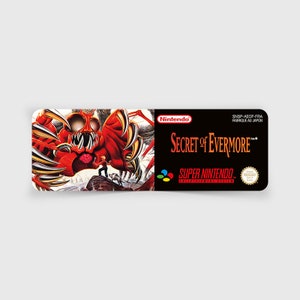 Puede incluir: Un cartucho de juego Super Nintendo para Secret of Evermore. La parte frontal del cartucho presenta un monstruo rojo con garras y dientes afilados. El título del juego está impreso en blanco sobre un fondo negro. El logotipo de Nintendo está en la esquina superior derecha.