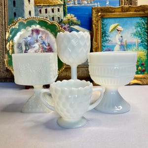 Puede incluir: Colección de cinco copas y tazas pequeñas de vidrio lechoso blanco con varios diseños en relieve. Las piezas incluyen una copa cuadrada, una copa con diseño de diamantes, una taza pequeña con asas y una copa con borde festoneado.
