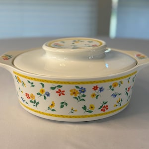 May include: A white ceramic casserole dish with a lid. The dish features a floral pattern of red, yellow, and blue flowers on a white base. The rim has a yellow border.