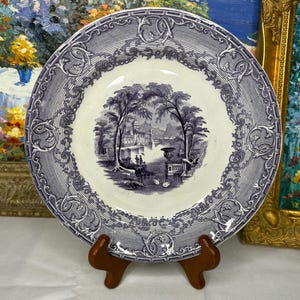 May include: A round, antique plate with a blue and white landscape design. The plate features a detailed border and a central scene of trees, buildings, and figures. The plate is displayed on a wooden stand.
