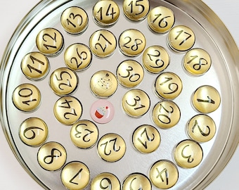 May include: A round metal tin filled with gold-colored round magnets with black numbers 1 through 31. Two small cupcake-themed magnets are also included.