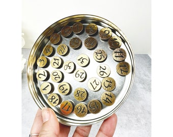 May include: A metal tin filled with round gold-colored tokens, each with a black number printed on it. The numbers range from 1 to 31.