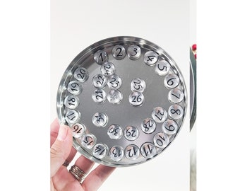 May include: A silver metal container with a circular top, holding 30 small silver circles with black numbers 1 through 30 printed on them.