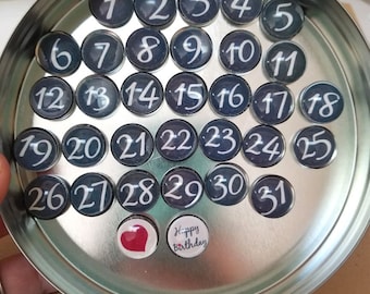 May include: A silver tin with a black and white heart and the words "Happy Birthday" on it. The tin contains 31 small round black and white magnets with white numbers 1 through 31.