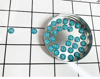 May include: A metal container with a white background holds a collection of teal-colored round discs with white numbers from 1 to 30. The discs are scattered around the container.